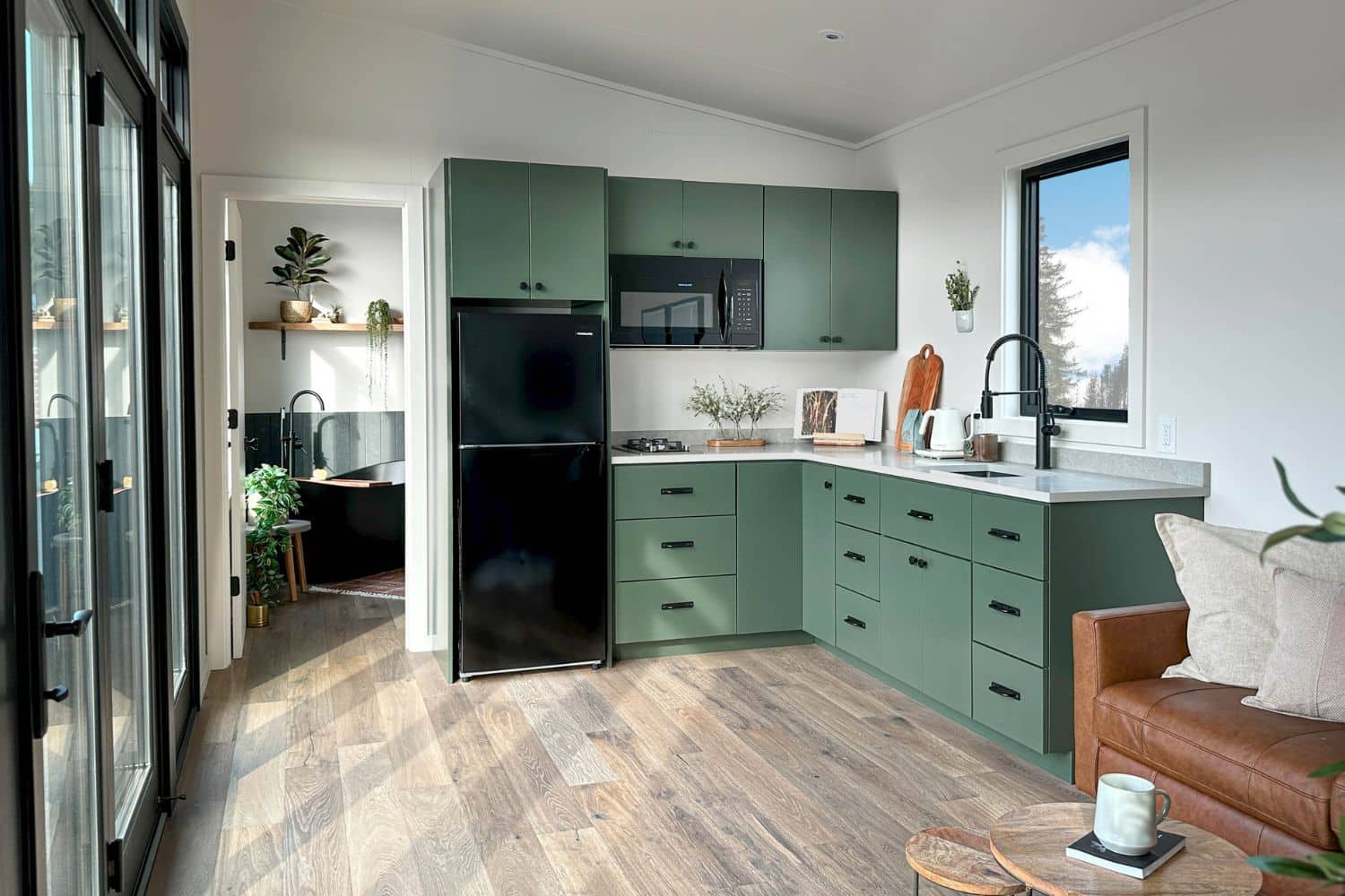 Kitchen in a tiny home with green cabinets. View out window and sliding glass doors on side.