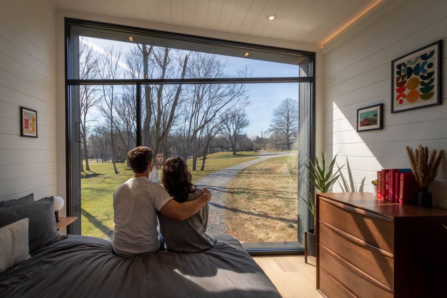 Pair sitting on bed in tiny home bedroom, facing window, looking out at trees and grass
