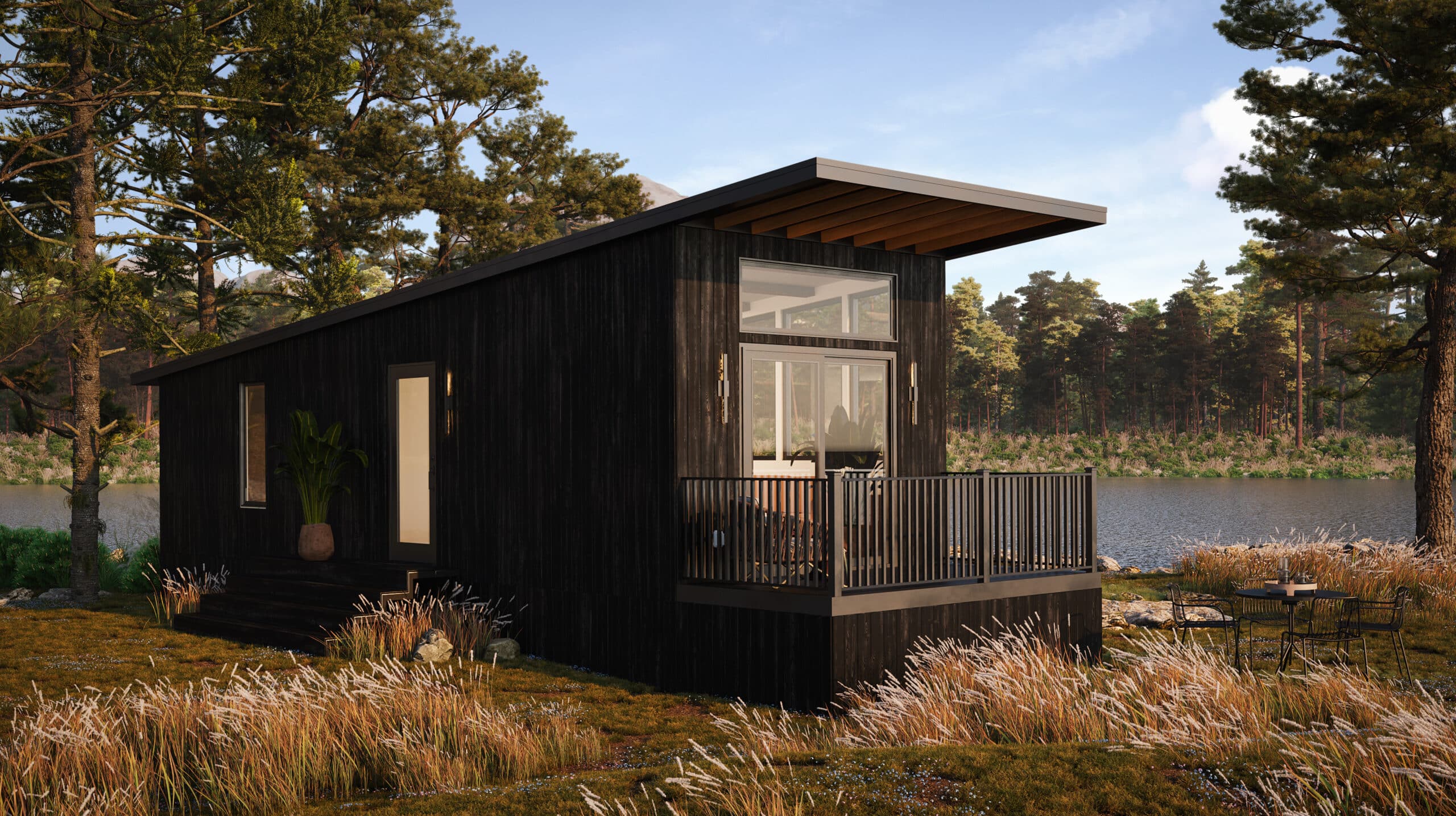 Modern black tiny home with large windows and a wooden deck, set by a peaceful lakeside surrounded by tall trees and grass.