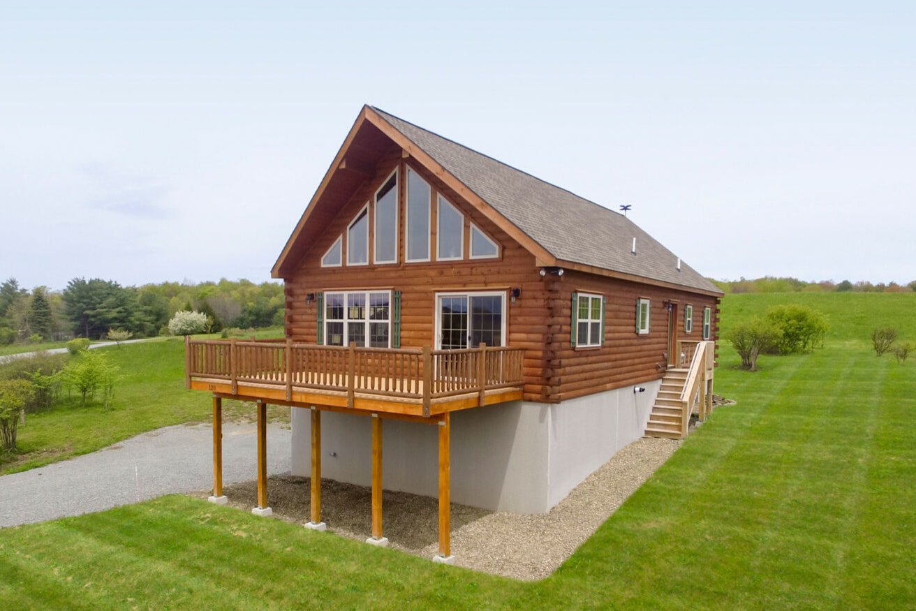 Chalet-style log cabin with high-pitched roof and raised deck in a grassy landscape.