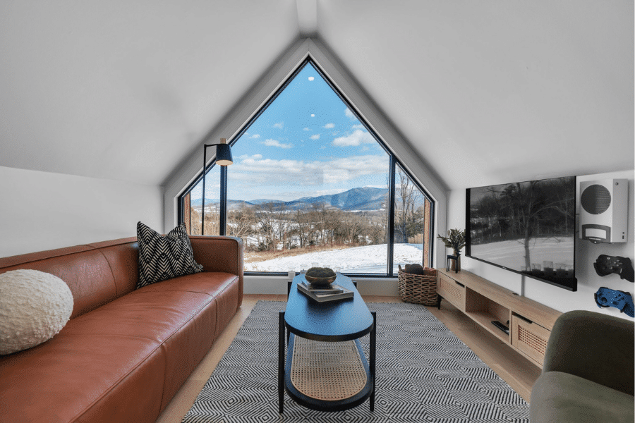 Interior view of a modern A-frame ADU living room with a leather sofa, geometric rug, coffee table, and entertainment setup, featuring a large triangular window framing a snowy mountain landscape.