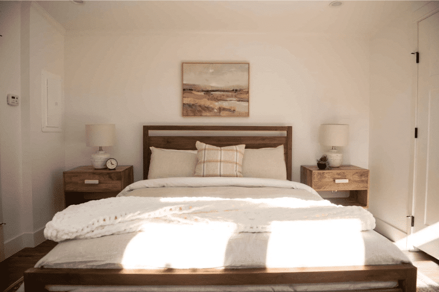 Cozy bedroom inside an ADU with white walls, natural wood bed frame and nightstands, beige bedding, and two lamps on either side of the bed.