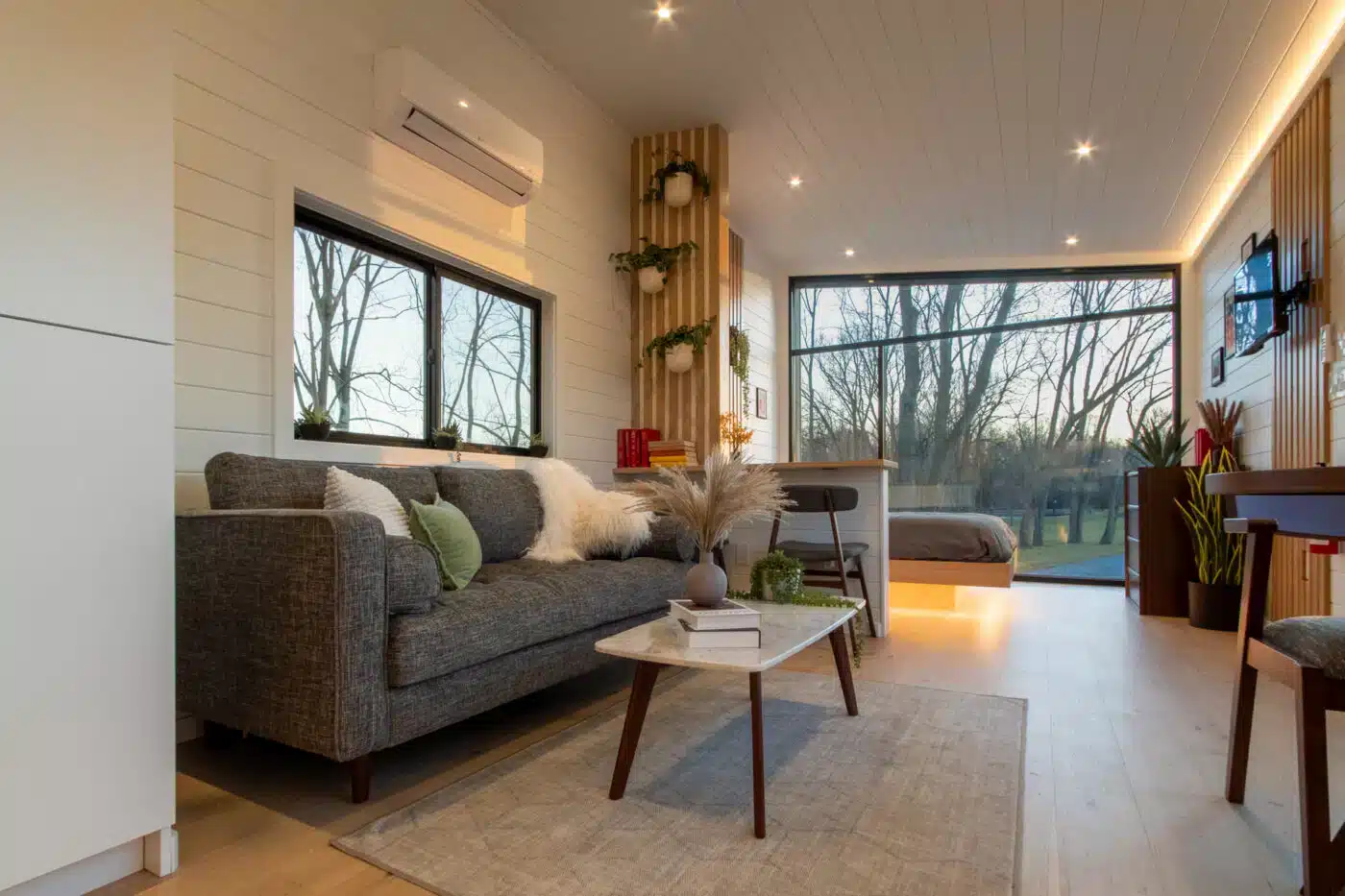 Cozy tiny home interior featuring a modern living room with a gray sofa, a coffee table, and a warm, natural color palette, leading to a bedroom with large windows offering scenic outdoor views.





