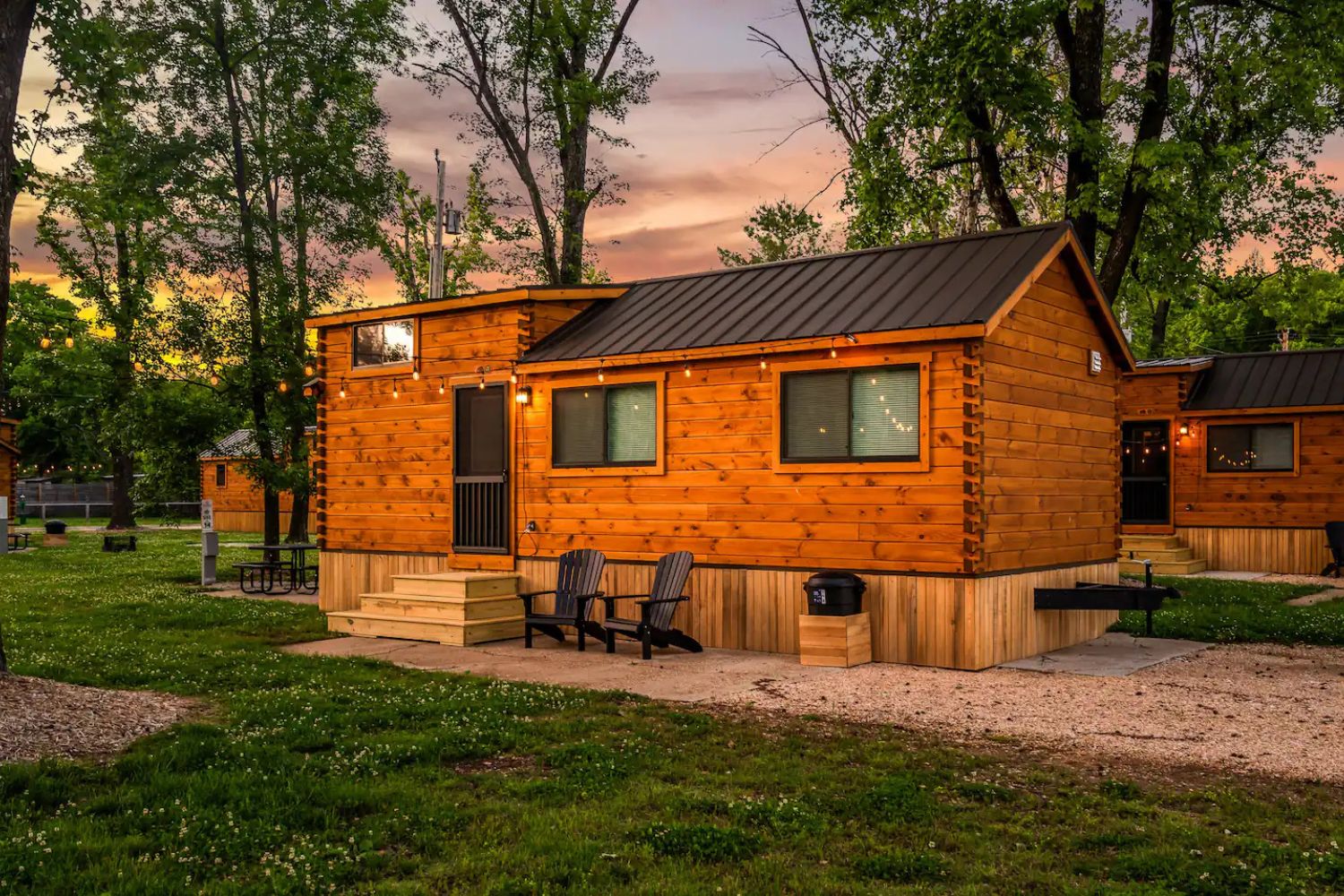 A charming park model home with rich wooden siding and a black metal roof is surrounded by lush greenery. The home features a small set of wooden steps leading to the front door, with warm outdoor string lights adding a cozy ambiance as the sun sets in the background. Two black Adirondack chairs are placed on a concrete patio, providing a relaxing spot to enjoy the peaceful surroundings. Additional amenities like a picnic table and neighboring cabins enhance the inviting atmosphere of this serene outdoor retreat.