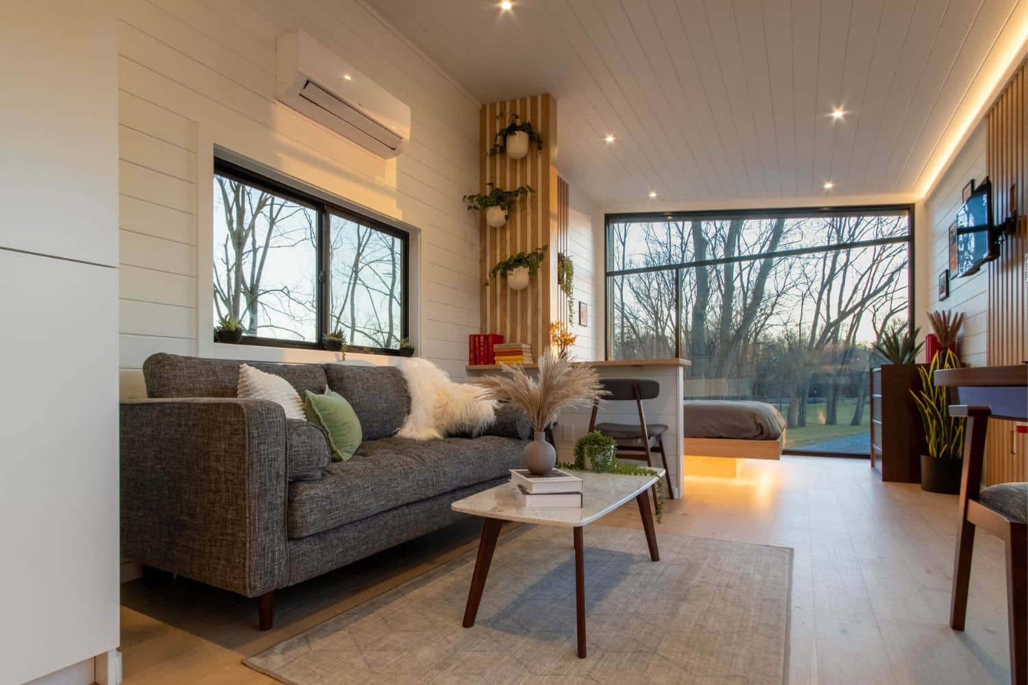 A bright and modern tiny home interior featuring a cozy gray sofa adorned with soft throw pillows and a blanket, complemented by a sleek marble-top coffee table holding decorative plants and books. The open floor plan leads to a compact dining area with stools and a neatly tucked bed with warm lighting underneath. Large windows provide expansive views of an outdoor wooded setting, allowing natural light to flood the space. Vertical wooden accents on the wall, adorned with hanging plants, add a touch of greenery and elegance. The minimalist design is enhanced by recessed ceiling lights and a neutral color palette, creating an inviting and serene atmosphere.