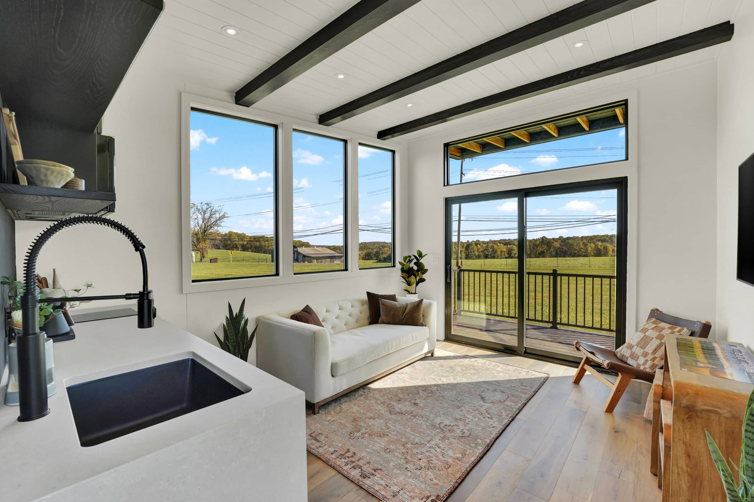 Bright and airy interior of a modern tiny home featuring large windows with scenic countryside views, a cozy white sofa, wooden flooring, and a sleek black kitchen sink in the foreground.